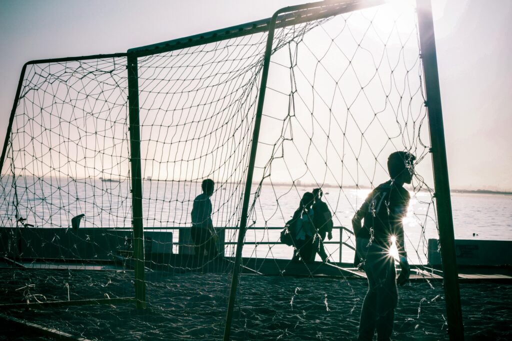 Silhouettes of people around a soccer goal on a sunny beach by the sea.